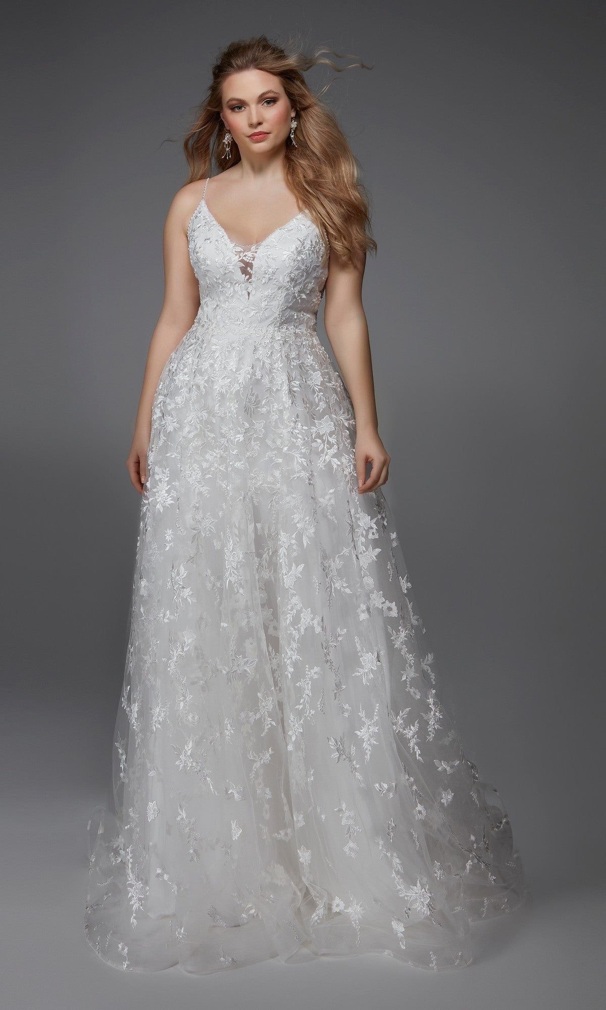 A woman wears the Alyce 7012 Long Formal Dress in ivory, featuring a sleeveless, floor-length lace design with floral appliques and an A-line silhouette, standing against a plain gray background.